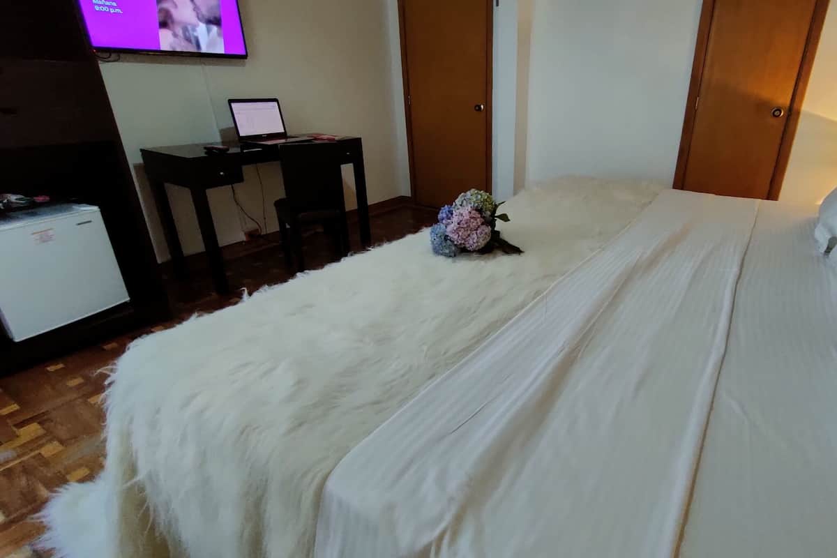 A hotel room with a large bed, a desk with a laptop, a television, and a small refrigerator.