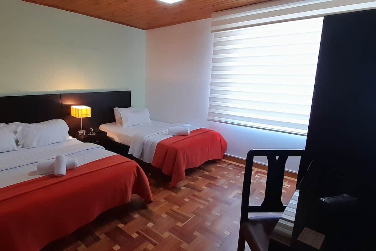 A hotel room with two beds, a wooden floor, a nightstand with a lamp, and a window with blinds.