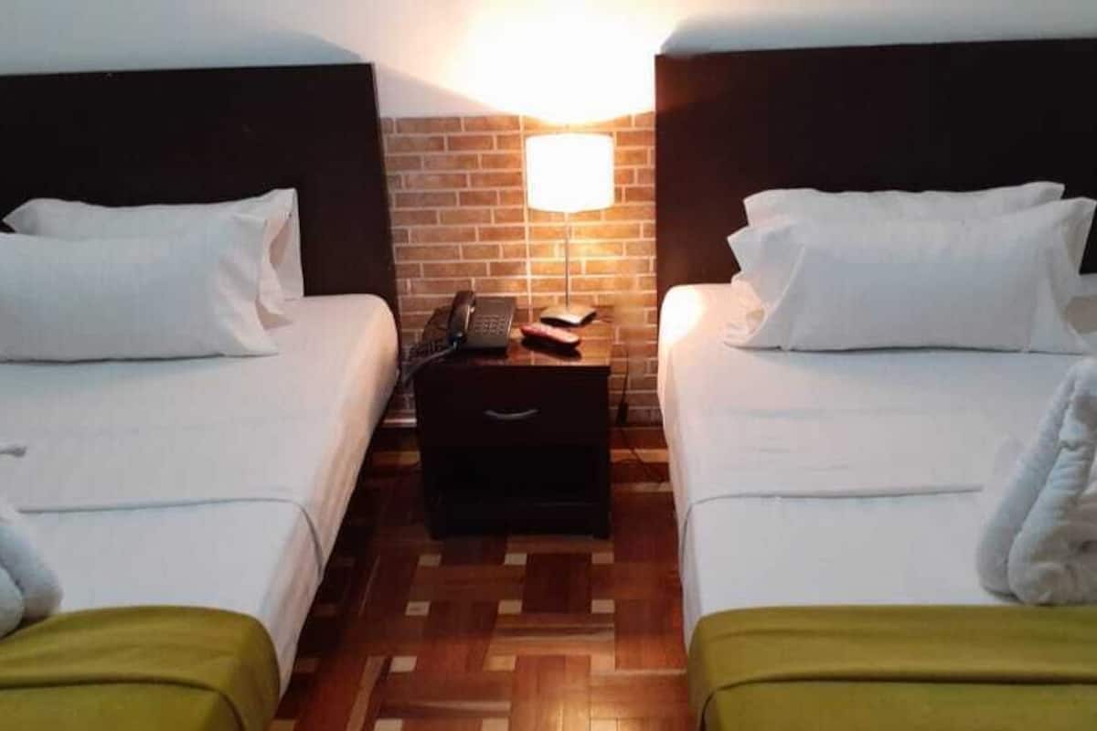 A hotel room with two beds, a nightstand with a lamp, and a brick-patterned wall.
