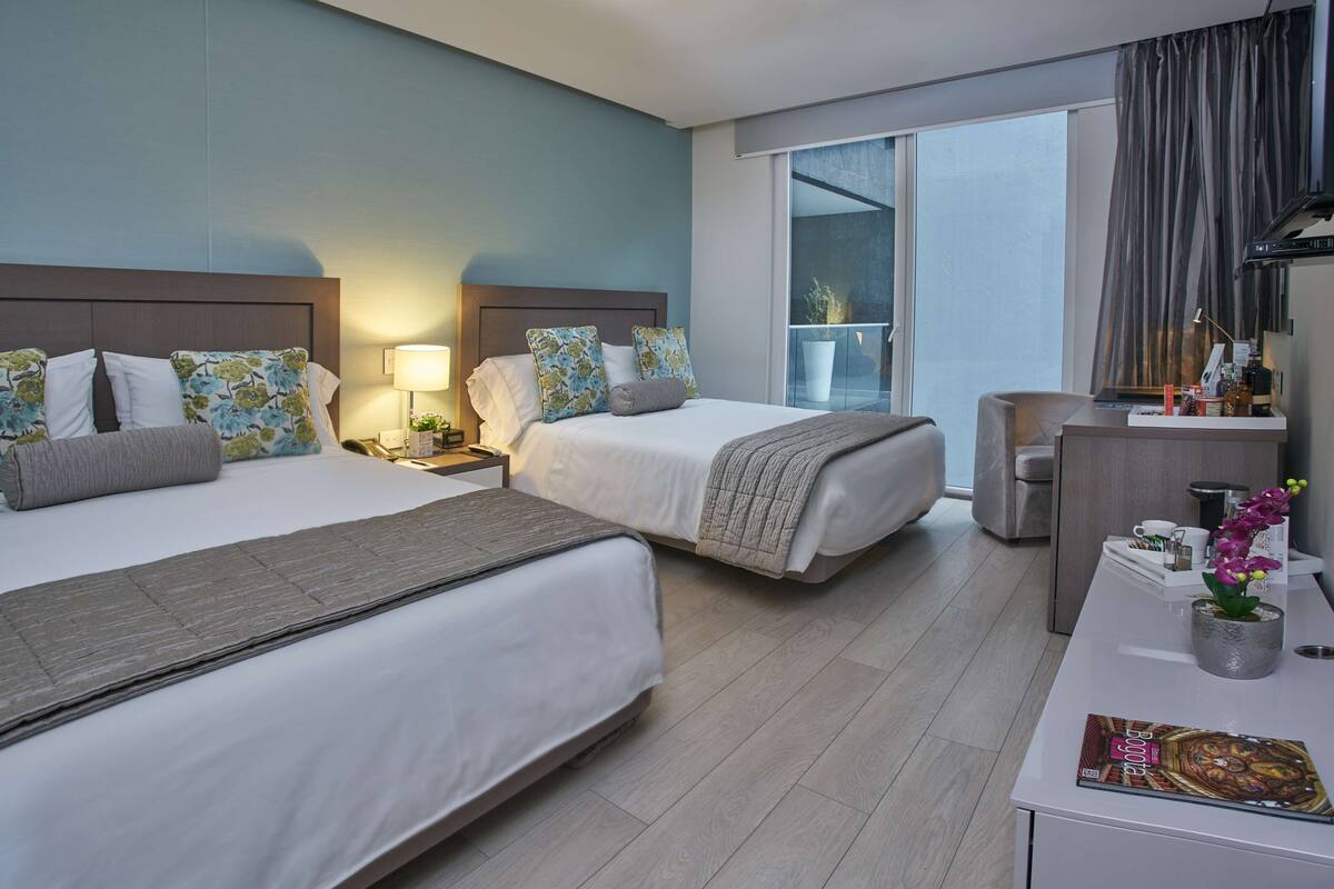 Habitación de hotel con dos camas, un escritorio, una silla, un balcón con vistas y un ventanal grande.