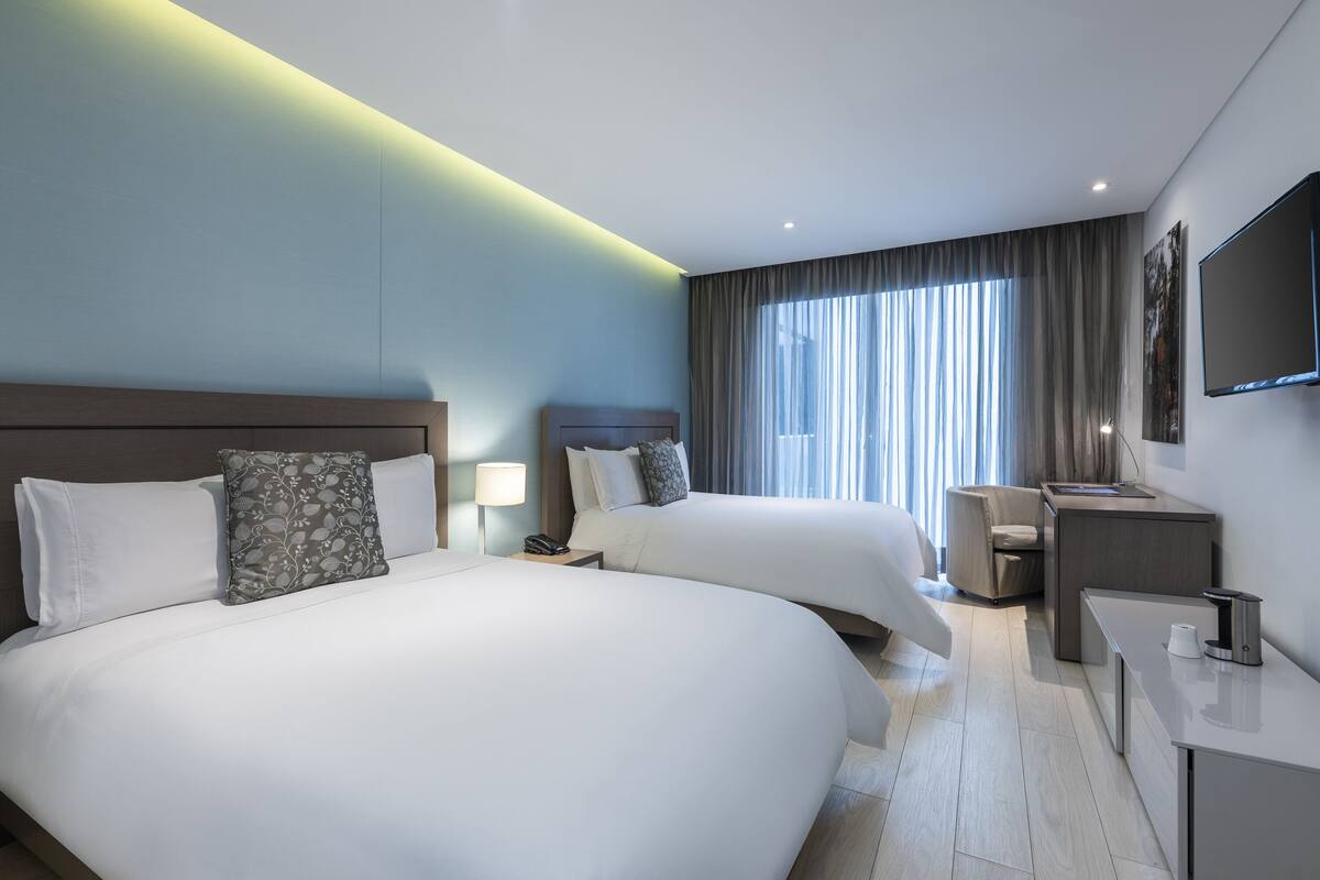 Una habitación de hotel moderna con dos camas grandes, un escritorio, una silla y un ventanal con cortinas.
