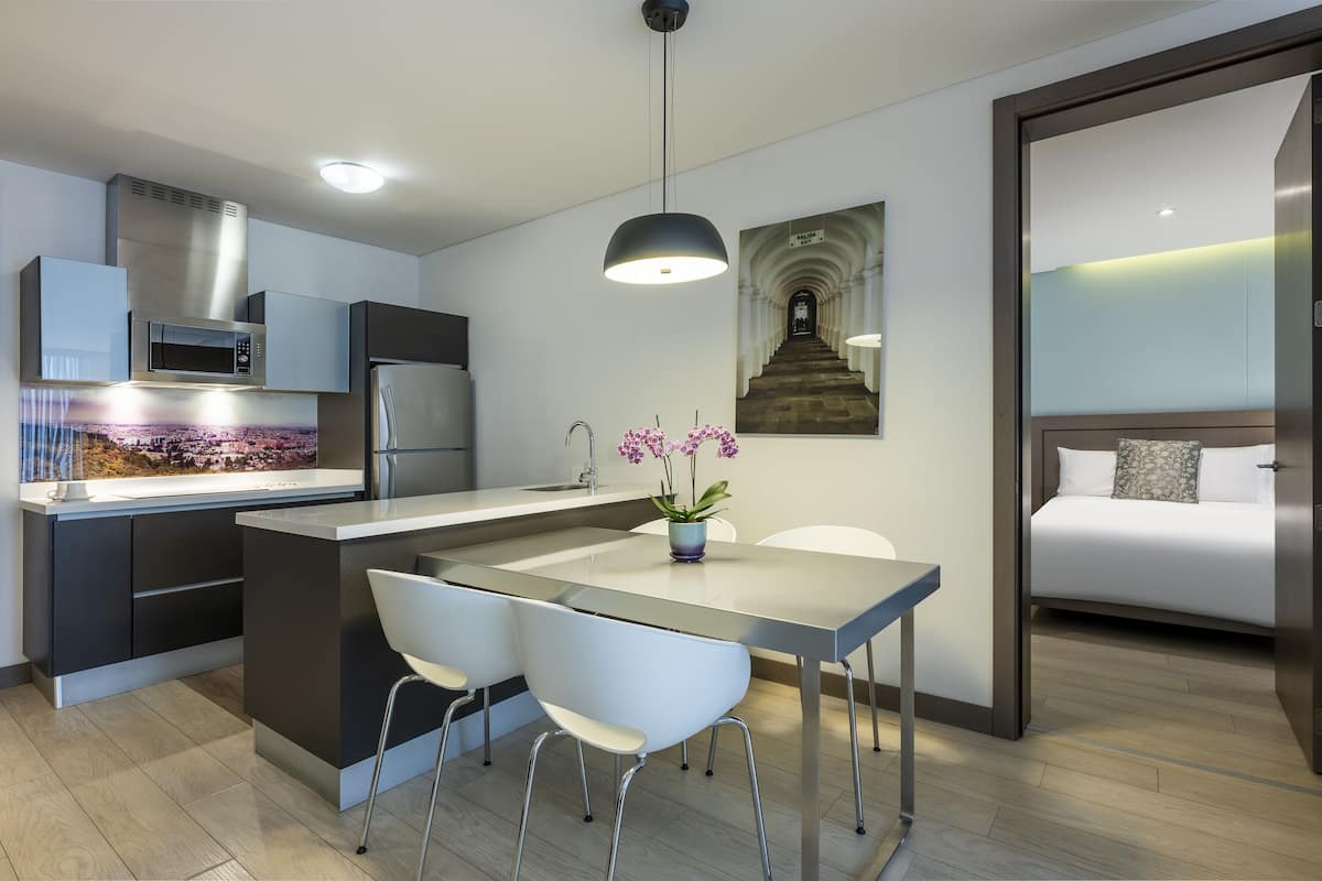 Una cocina moderna con una isla central, una zona de comedor con dos sillas, una lámpara colgante y una cama con vista a una escalera.