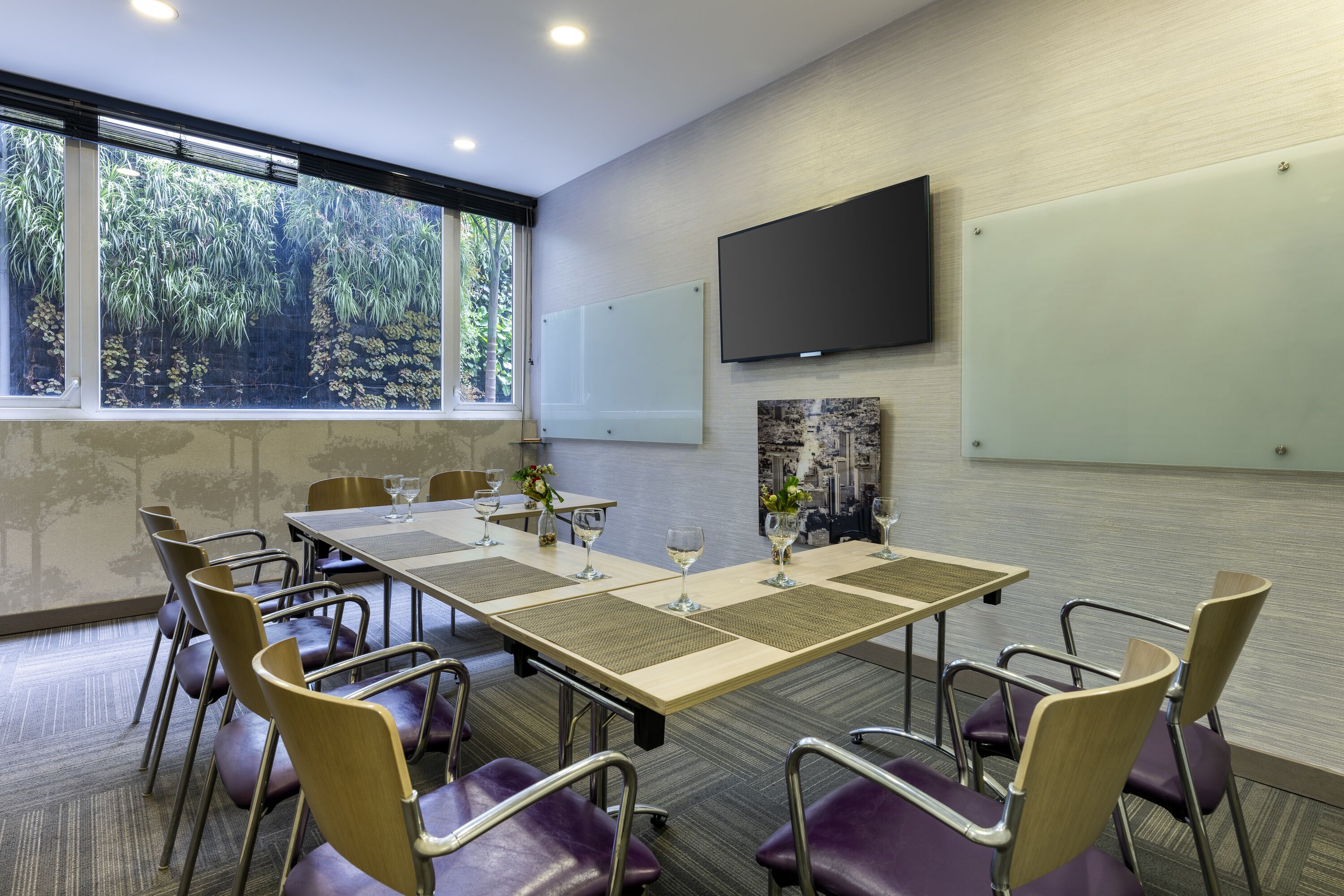 A conference room with a long table, chairs, a TV screen, and a window with a view of trees.