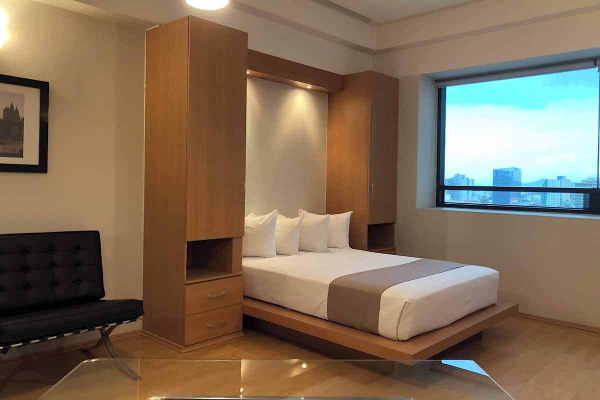 A modern bedroom with a bed, a wooden wardrobe, a black sofa, and a city view through a window.