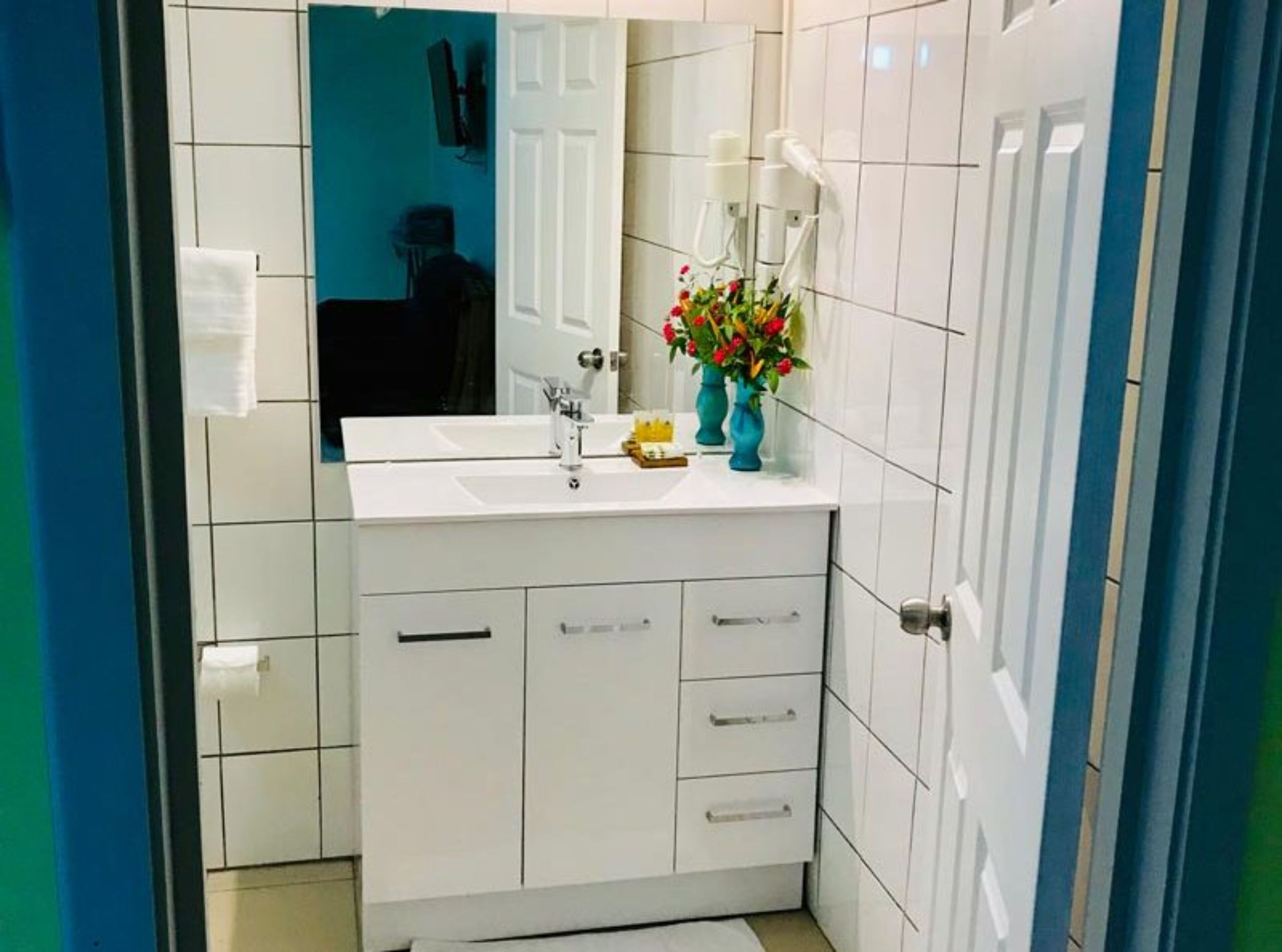 A bathroom with white tiled walls, a white sink, and a mirror. There are two white cabinets with drawers and a small shelf with a vase of flowers.