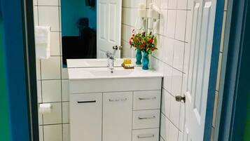 A bathroom with white tiled walls, a white sink, and a mirror. There are two white cabinets with drawers and a small shelf with a vase of flowers.