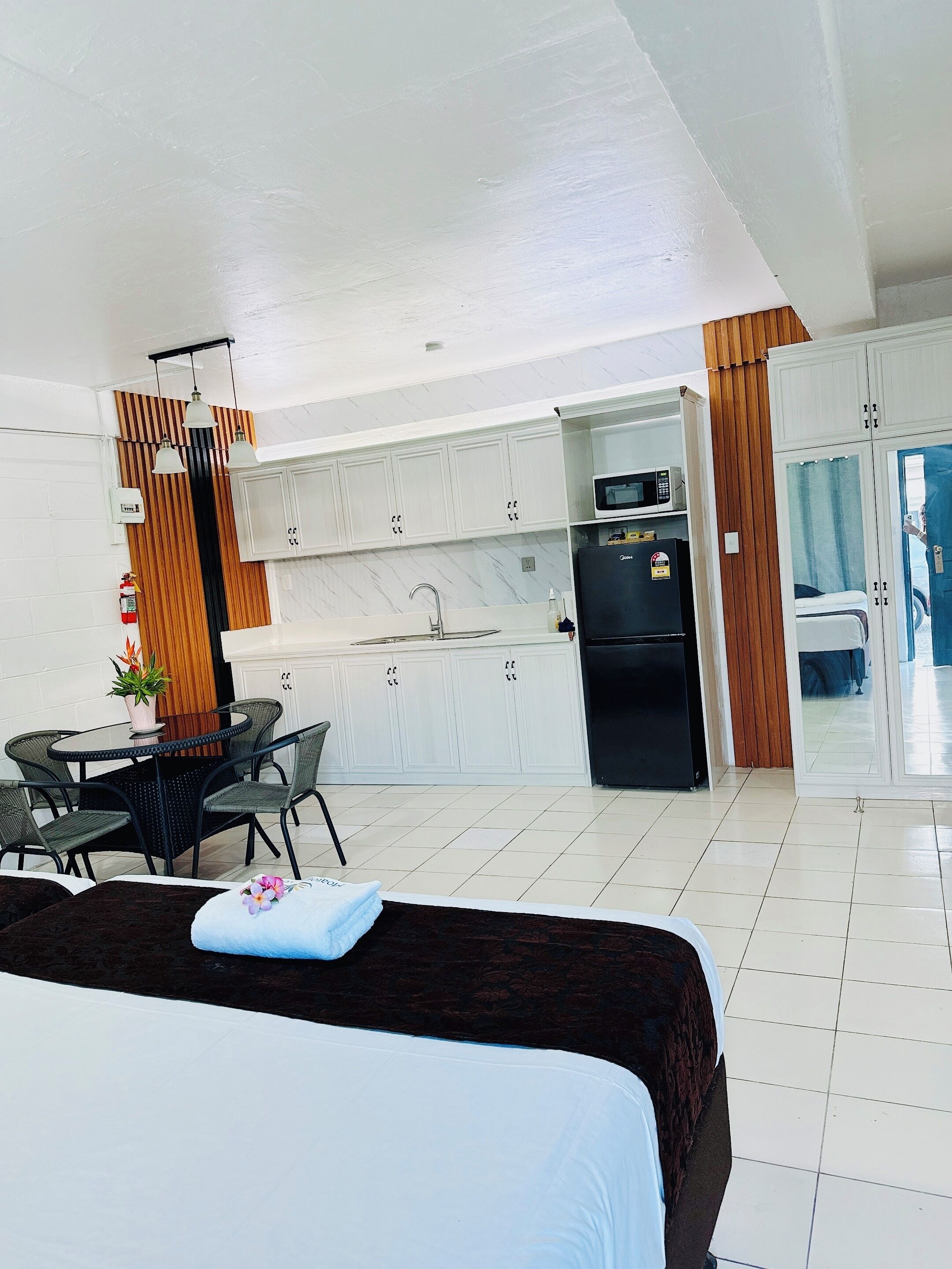 Habitación de hotel moderna con cama, un pequeño comedor con mesa y sillas, una kitchenette con armarios blancos y vistas a través de una puerta.