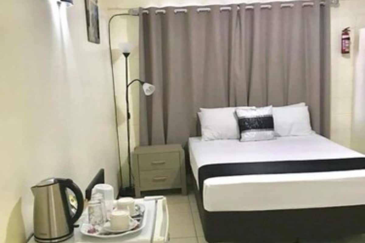 A hotel room with a bed, bedside table, lamp, and a tray with a kettle and cups.