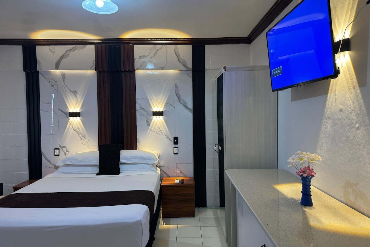 Habitación de hotel con cama, mesita de noche, televisor de pared y un jarrón con flores.