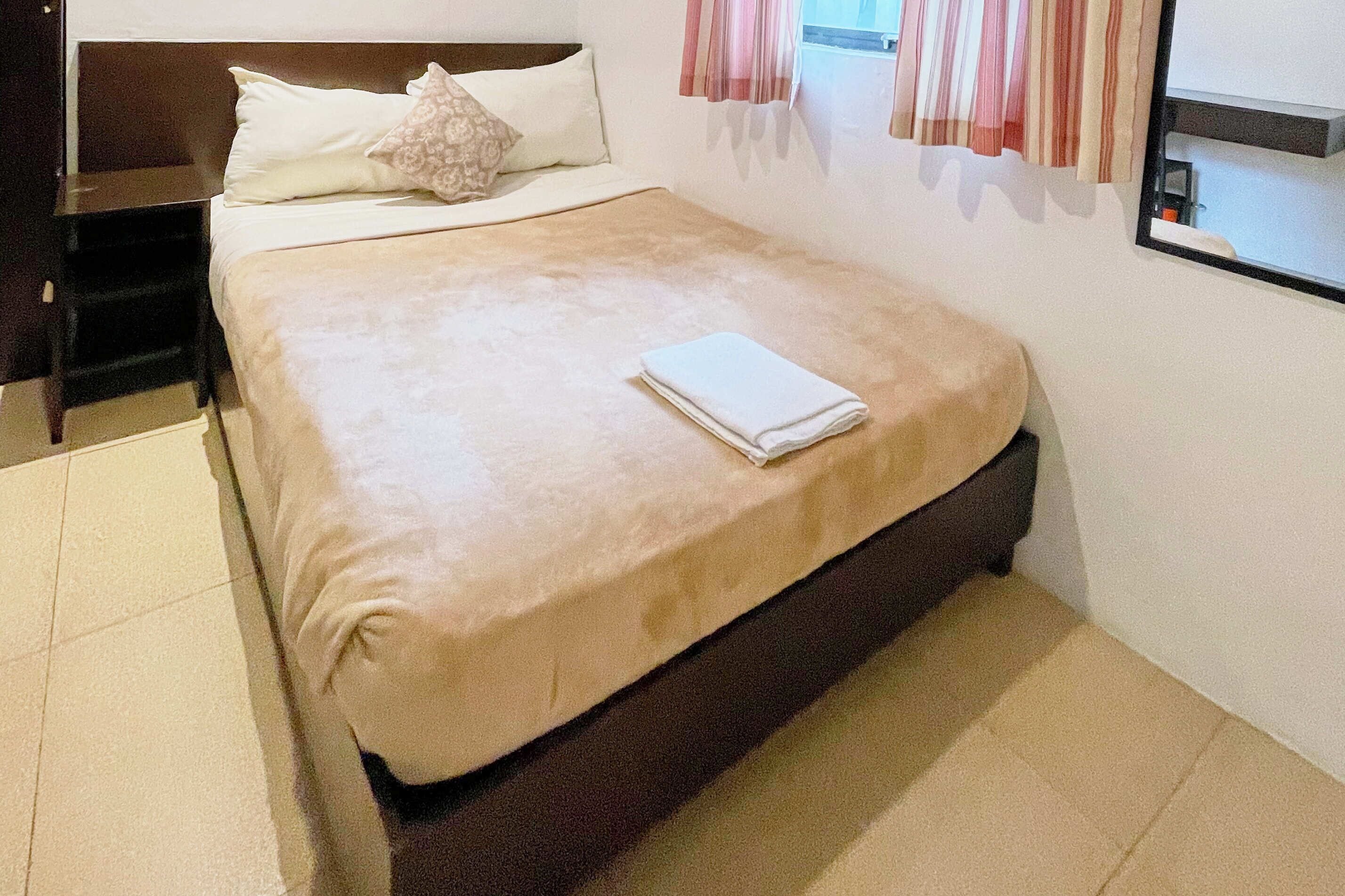 A neatly made bed with a folded white towel, a wooden nightstand, and a window with curtains.
