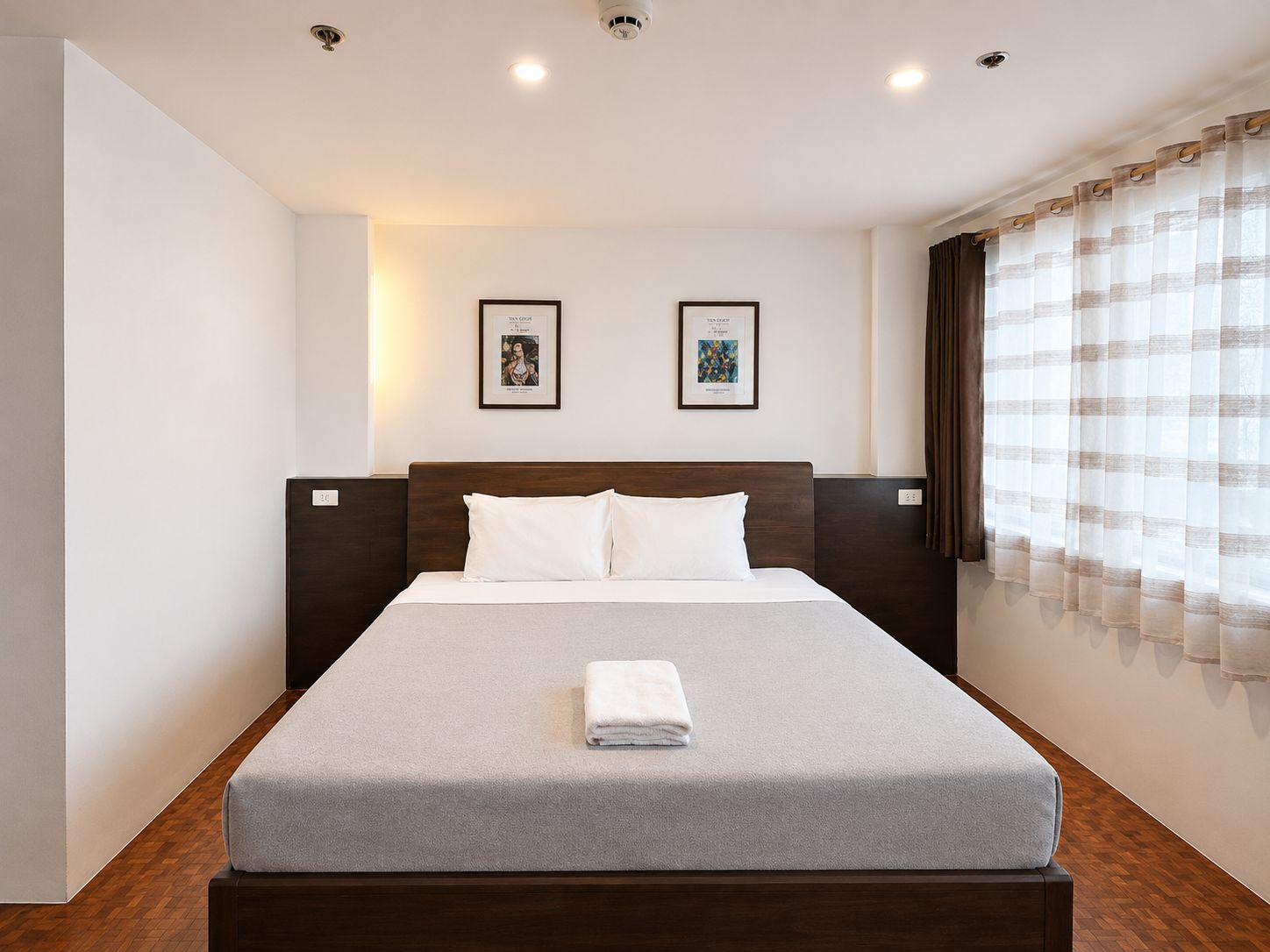 Habitación de hotel con una cama, dos cuadros en la pared, una ventana con cortinas y suelo de madera.