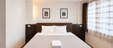 Habitación de hotel con una cama, dos cuadros en la pared, una ventana con cortinas y suelo de madera.