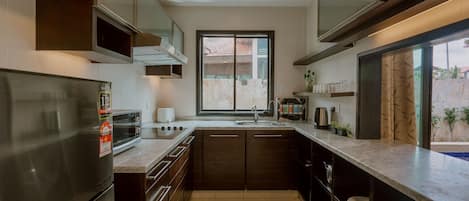 A modern kitchen with a stainless steel refrigerator, a built-in oven, a microwave, and a sink. There is a window with a view of a balcony and a door leading to another room.
