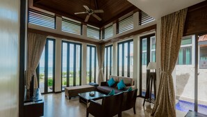 A spacious living room with a sofa, coffee table, and a ceiling fan. Large windows provide a view of the outdoors. - Borneo Beach Villa & Suite (Kota Kinabalu)