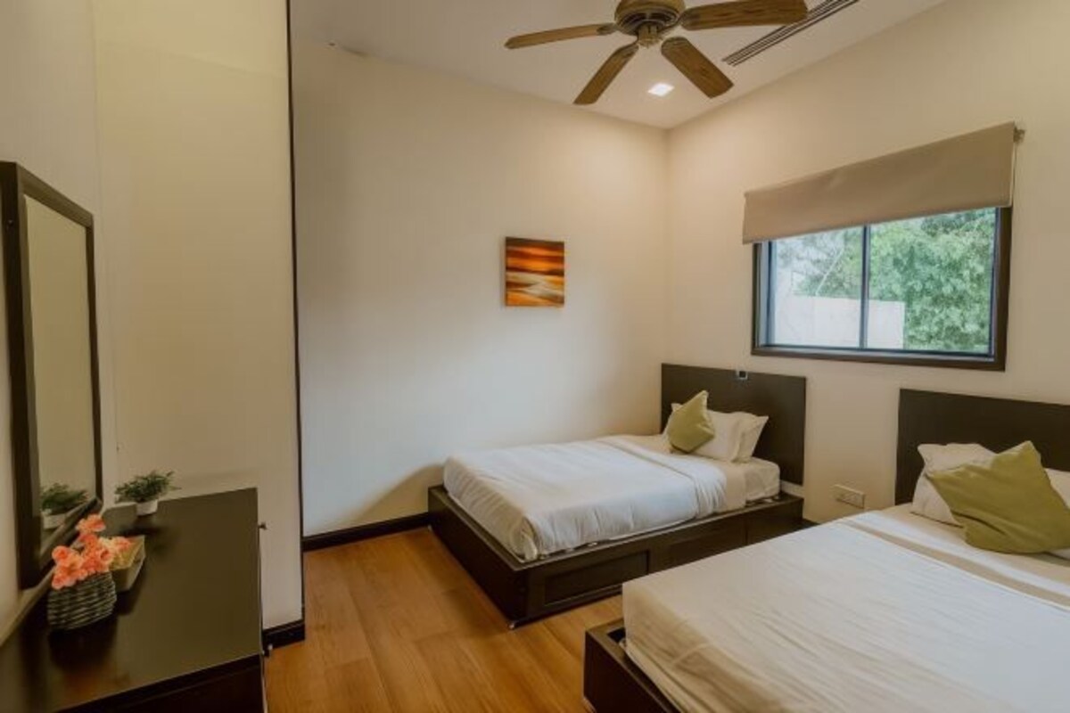 A bedroom with two beds, a ceiling fan, a window with a view, and a painting on the wall.