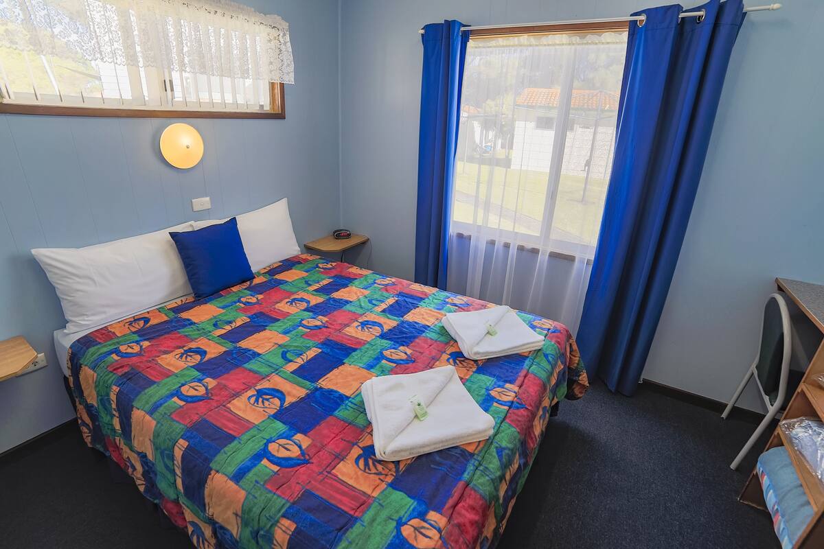 A bedroom with a bed, a nightstand, a desk, and a window with curtains.