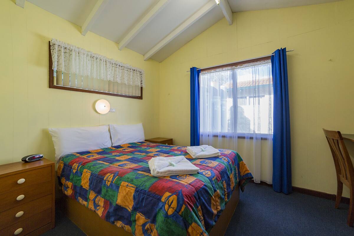 A hotel room with a bed, a nightstand, a desk, and a window with blue curtains.