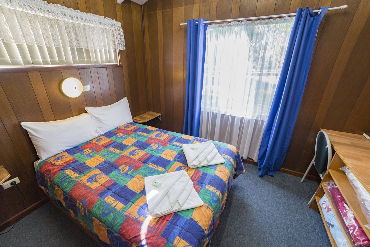 A hotel room with a wooden paneled wall, a bed with a colorful quilt, a small bedside table, a nightstand, a desk with a chair, and a window with blue curtains.