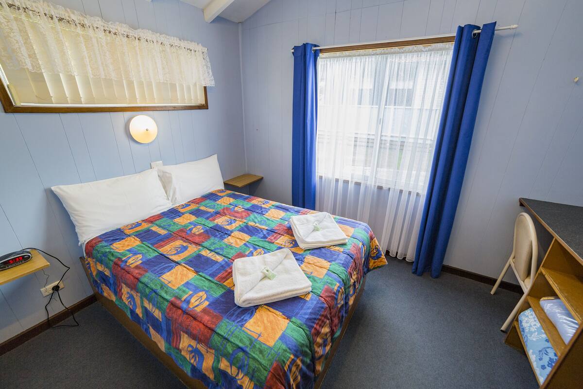 A room with a bed, a desk, and a chair. There is a window with curtains, a wall-mounted lamp, and a power outlet.