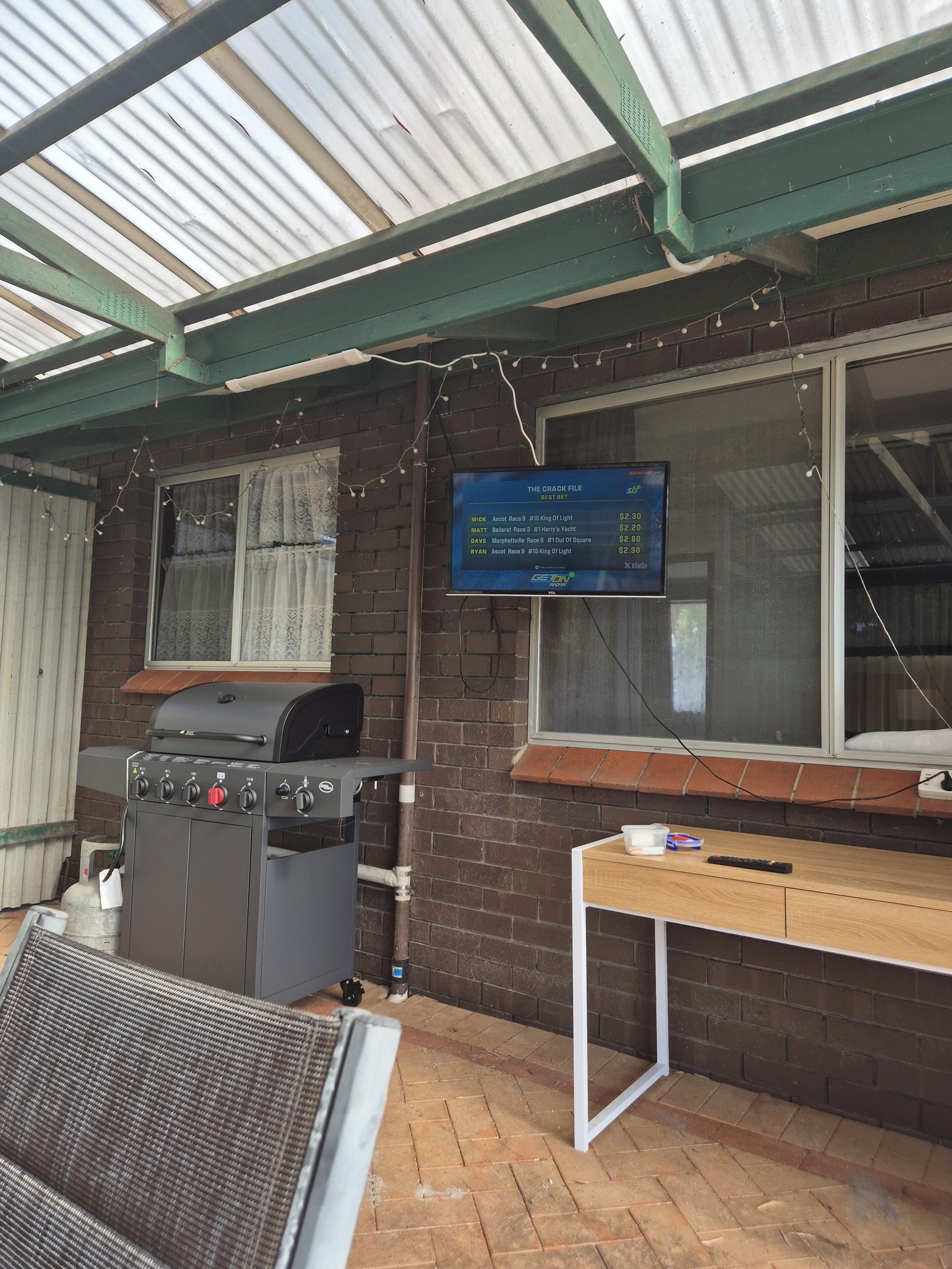 A covered patio area with a grill, a table with a remote control, and a screen displaying a race list.
