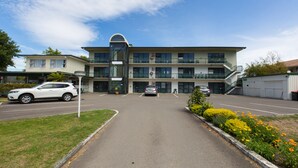A multi-story hotel with a central glass elevator, a parking lot with several cars, and a landscaped garden with yellow flowers. - Gateway International Motel (Rotorua)