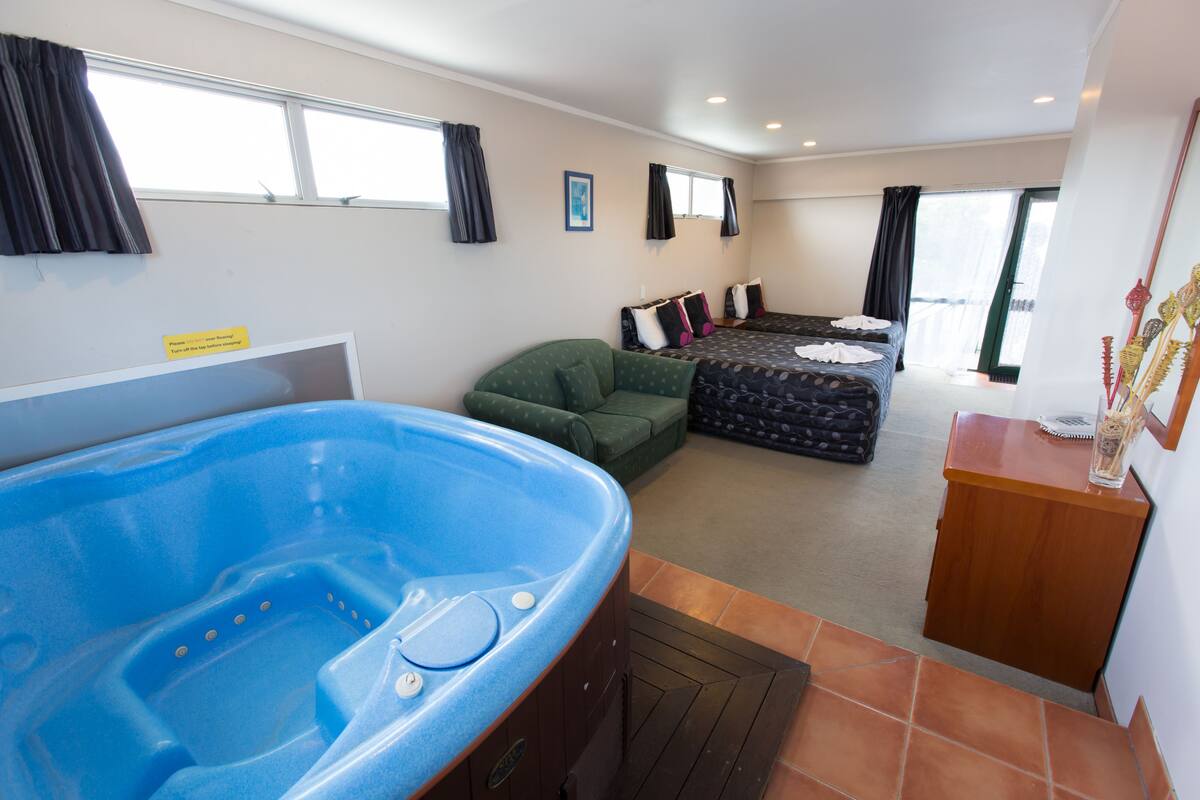 A room with a large blue hot tub, a green sofa, a bed with multiple pillows, and a wooden side table with a bottle.