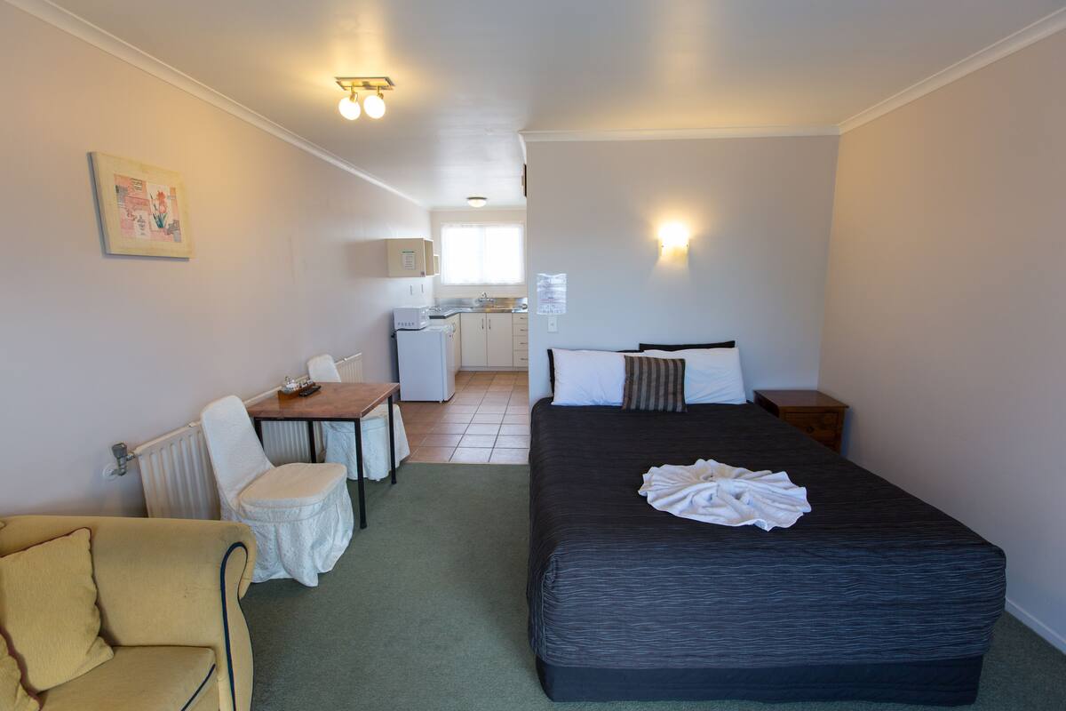 A compact hotel room with a bed, a small desk, a chair, and a kitchenette in the background.