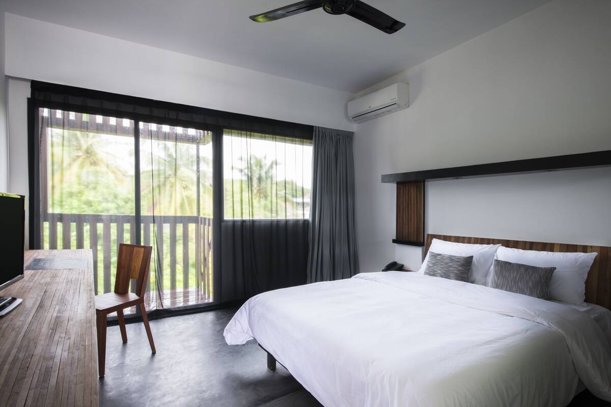 Ein Hotelzimmer mit einem Bett, einem Schreibtisch, einem Sessel, einem Deckenventilator und einem großen Fenster mit Vorhängen.