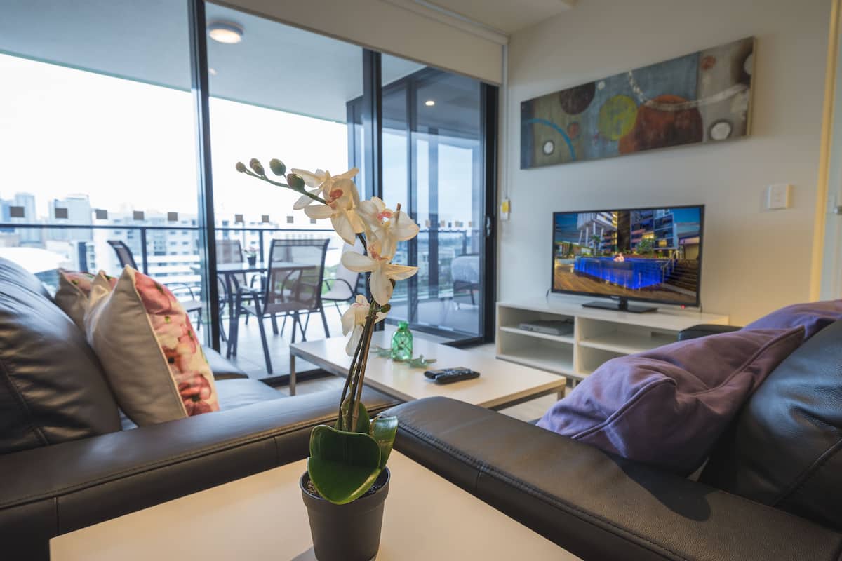 A modern living room with a sofa, a coffee table, a potted orchid, a television, and a balcony with a view of the city.