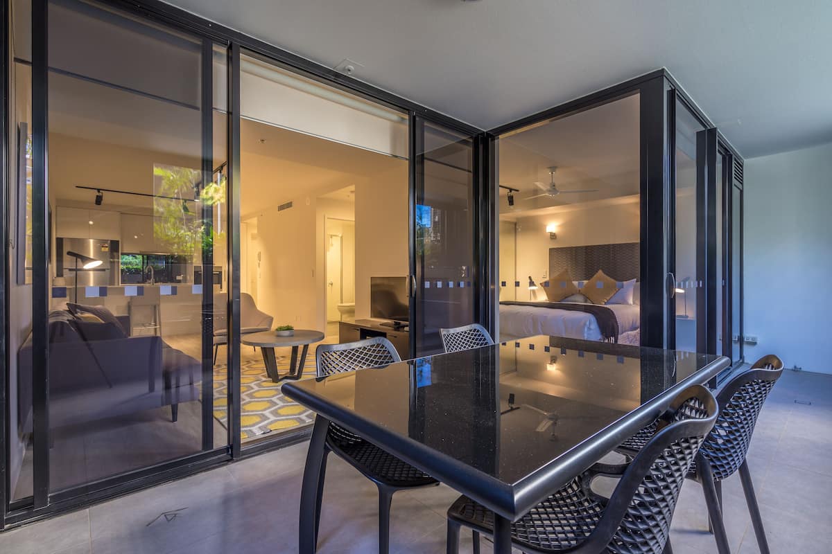 A modern hotel room with a glass wall, a dining area with a glass table and chairs, and a visible kitchen area.