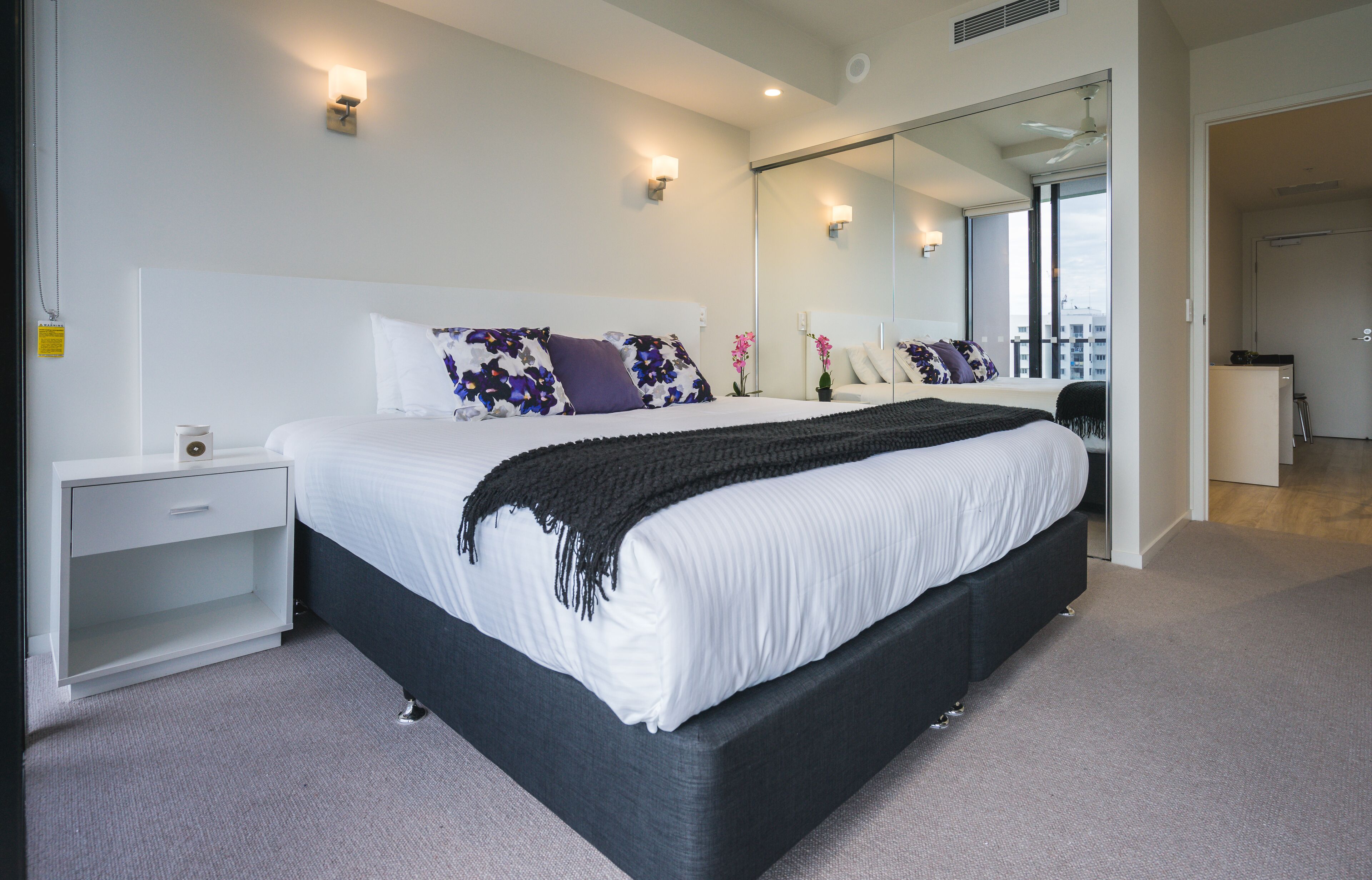 Un dormitorio moderno con una cama grande, mesitas de noche, un espejo y vistas a la ciudad.
