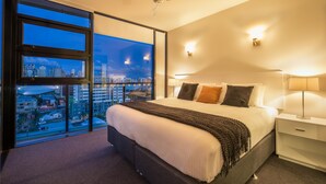 Une chambre d’hôtel moderne dotée d’un grand lit, offrant une vue sur la ville grâce à de larges fenêtres du sol au plafond, d’un ventilateur de plafond et d’une table de chevet avec une lampe.
