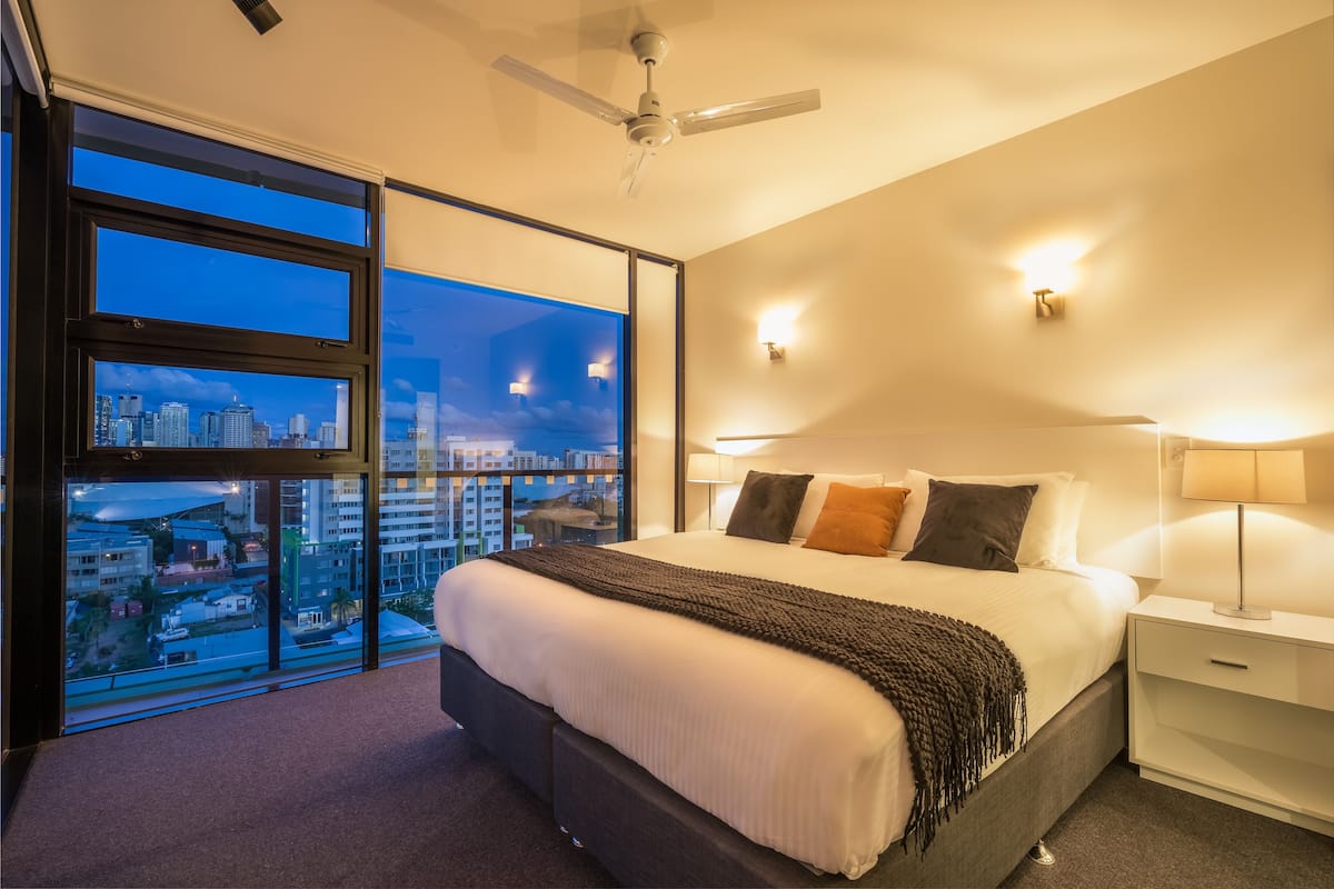A modern hotel room with a large bed, a city view through floor-to-ceiling windows, a ceiling fan, and a nightstand with a lamp.