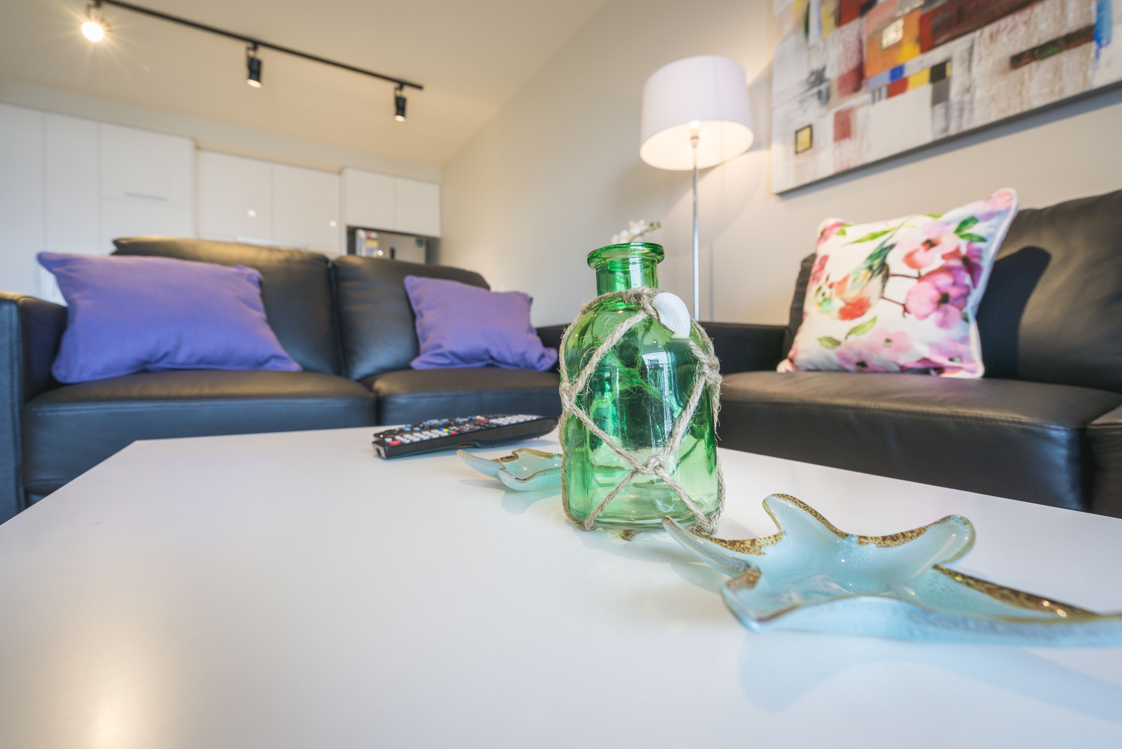 Una sala de estar con una mesa de centro blanca, una botella verde y una decoración con estrella de mar. Hay un control remoto en la mesa, y un sofá con cojines morados y estampados florales al fondo.