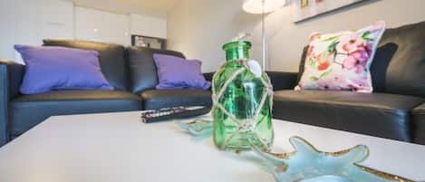 Una sala de estar con una mesa de centro blanca, una botella verde y una decoración con estrella de mar. Hay un control remoto en la mesa, y un sofá con cojines morados y estampados florales al fondo.