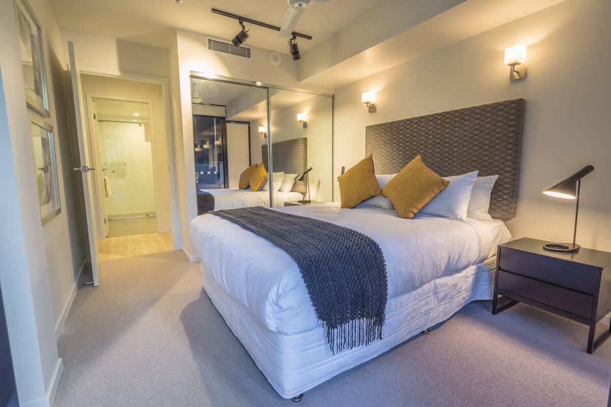 A modern hotel room with a large bed, a bedside table, a nightstand, a mirror, and a ceiling light.