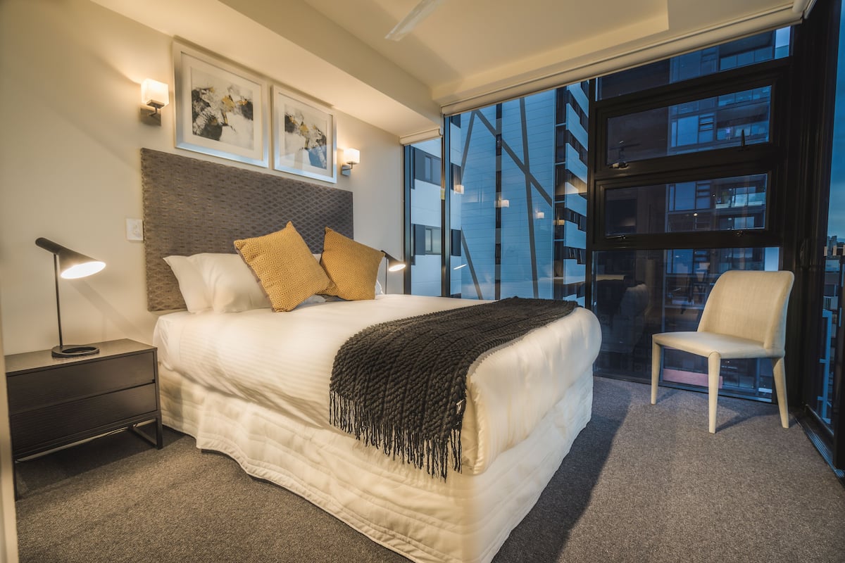 A modern hotel room with a large bed, a nightstand, a chair, and a view of a building through a glass door.