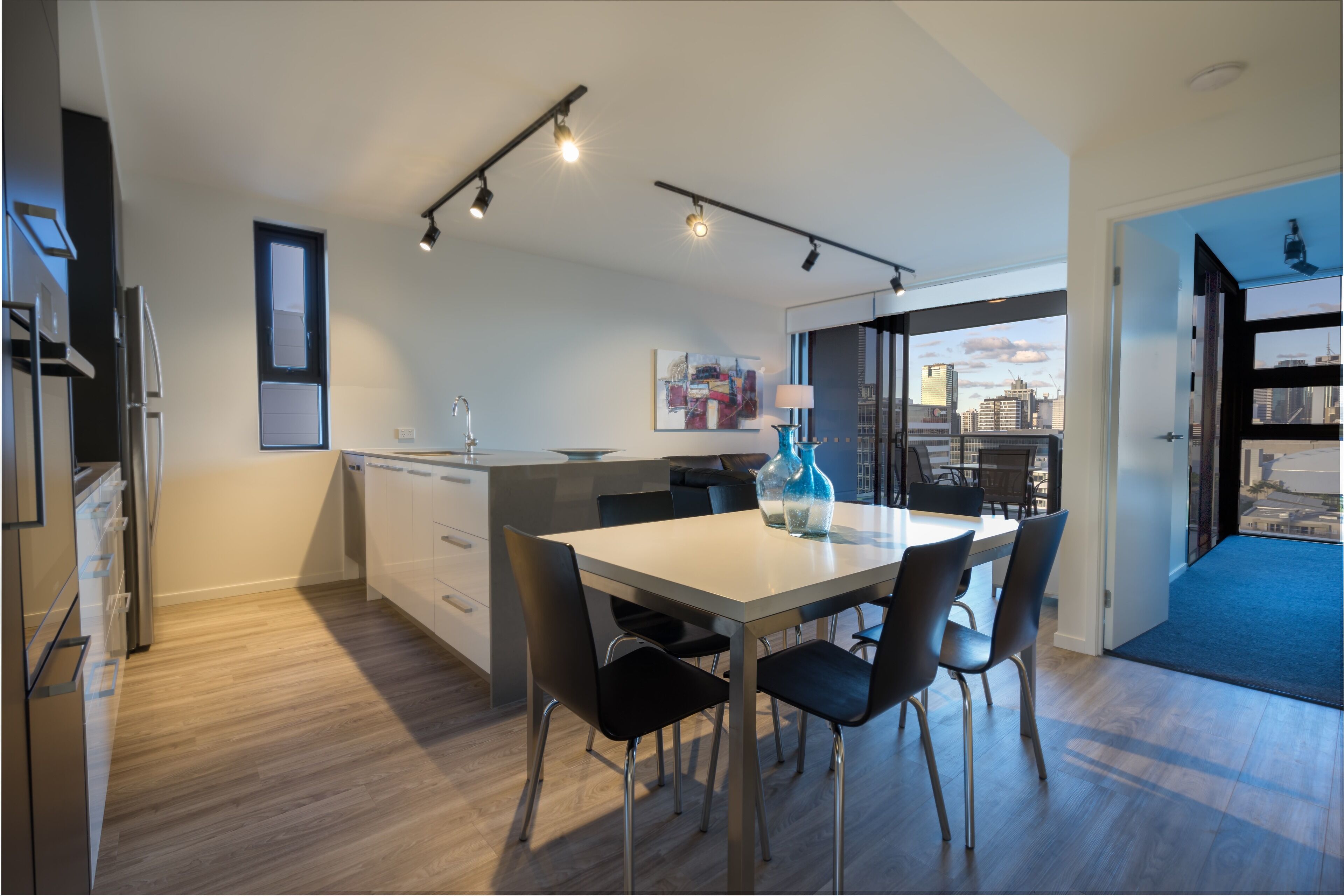 A modern kitchen with a dining area, featuring a large island, black chairs, and a view of the city through large windows.