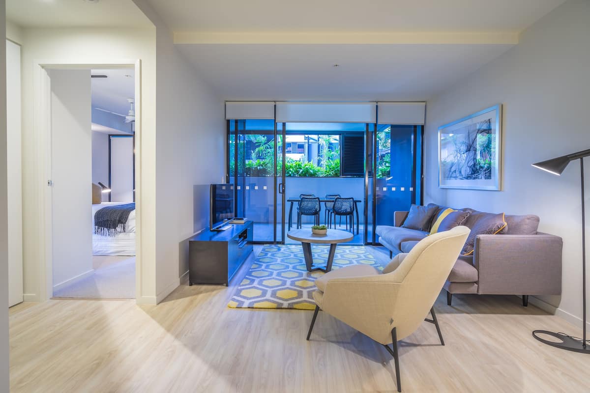 A modern living room with a sofa, armchair, and a small table with a plant. There is a balcony with a view of greenery and a door leading to another room.