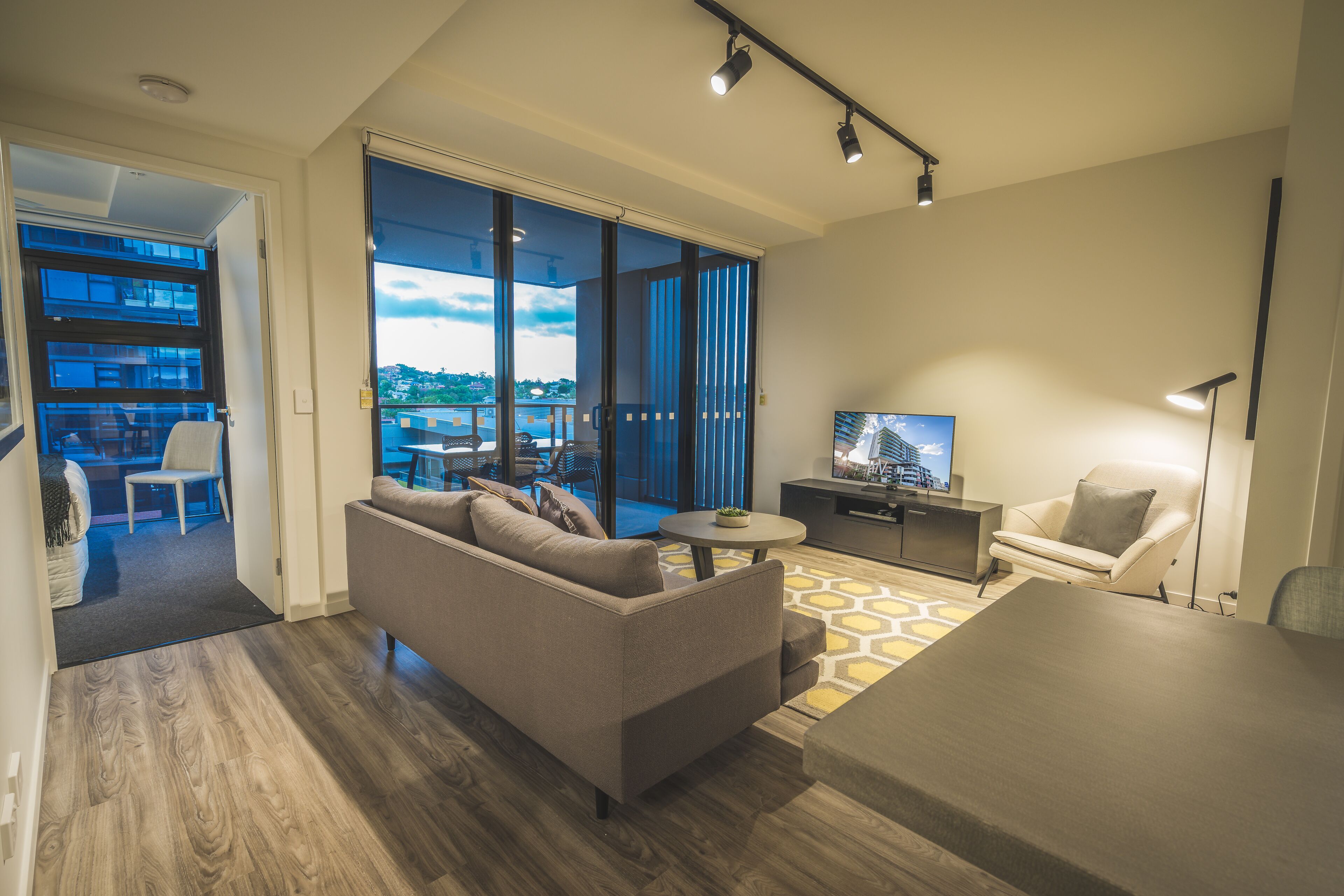 Ein modernes Wohnzimmer mit einer Couch, einem Couchtisch, einem Fernseher und einem Sessel. Ein großes Fenster bietet Blick auf die Stadt.