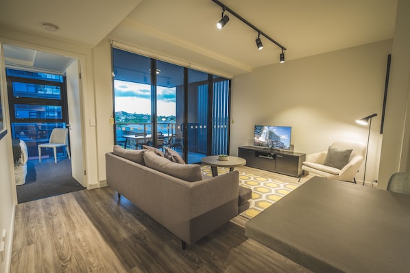 A modern living room with a sofa, a coffee table, a TV, and a chair. There is a large window with a view of the city.