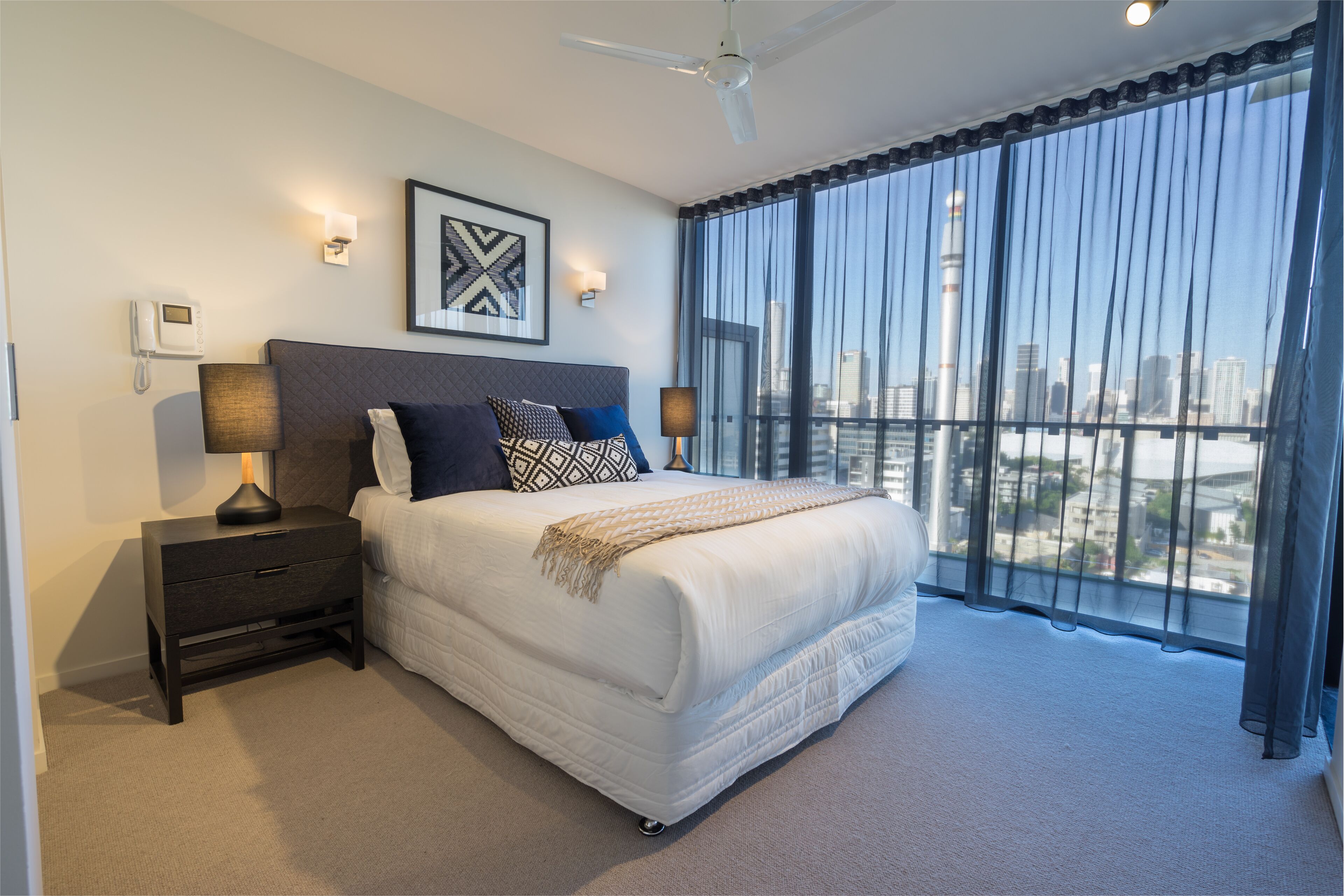 Une chambre d’hôtel moderne avec un grand lit, une table de chevet avec une lampe, un téléphone et une vue sur la ville à travers de fines rideaux.