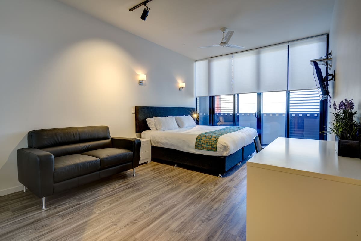 A modern hotel room with a bed, a sofa, a desk, and a large window offering a city view.