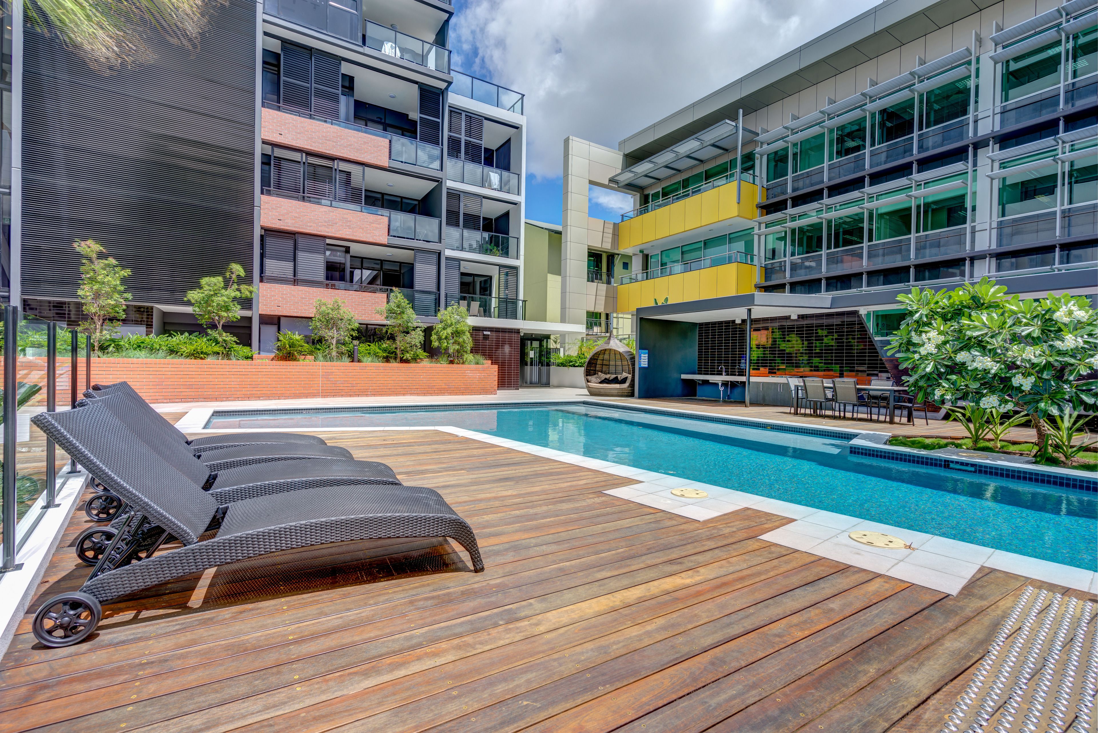Un área de piscina moderna con sillones de descanso, una terraza de madera y un edificio con varios balcones y ventanas amplias.