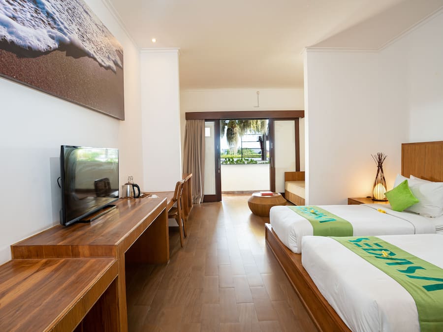 A hotel room with a wooden desk, a television, two beds with green bedspreads, a large painting of a beach, and a view of the outdoors through a glass door.