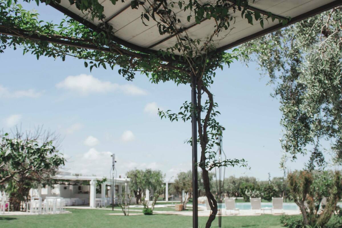 An outdoor patio with a table and benches, surrounded by greenery and a view of a building in the background.