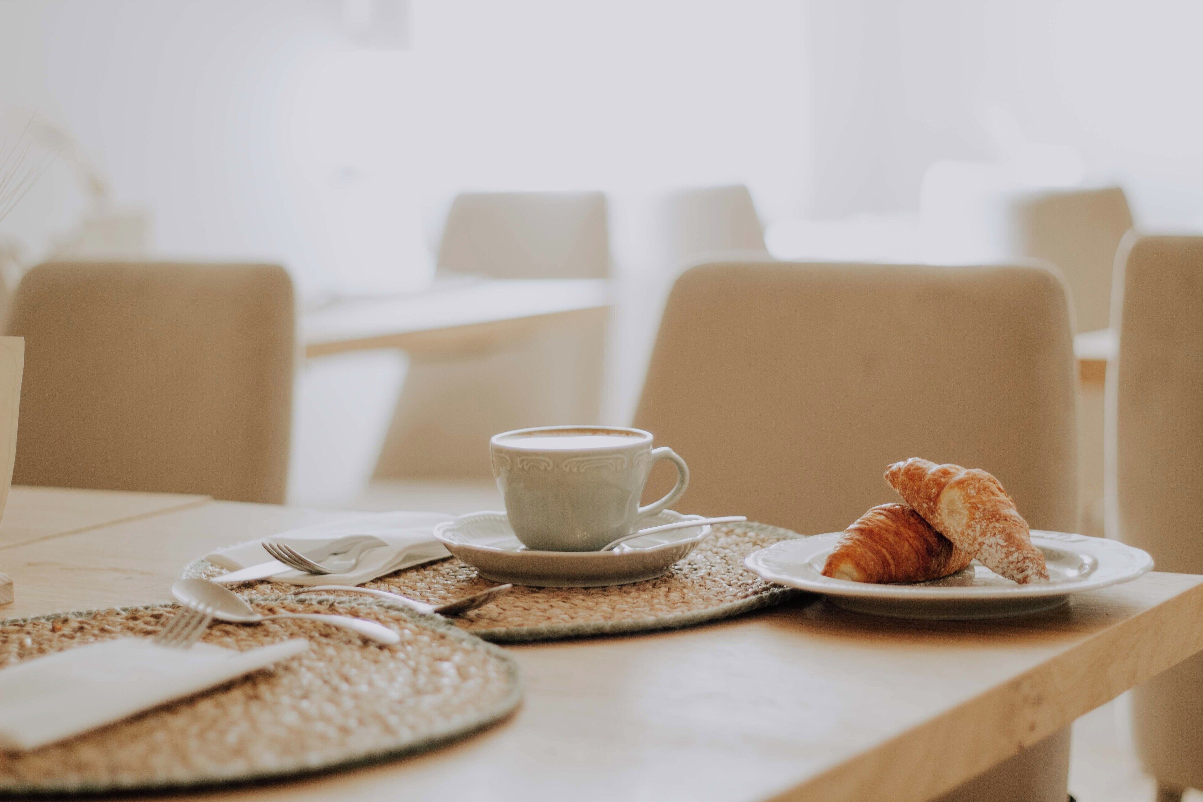 Ein Tisch gedeckt mit einer Kaffeetasse, einem Croissant und einem Teller Croissants, einer Tischmatte und einem Servietttuch.