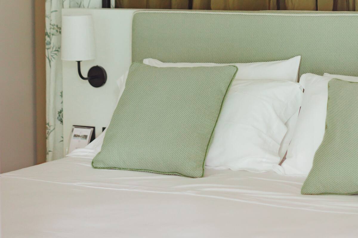 A hotel room with a bed, green pillows, a white headboard, and a curtain with a floral pattern.