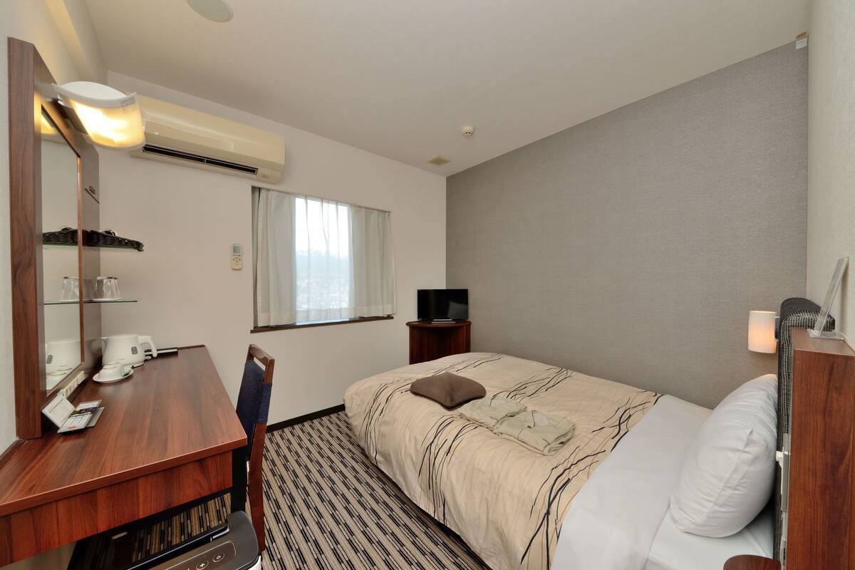 A hotel room with a bed, a desk with a chair, a television, and an air conditioning unit.