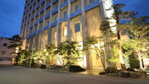 Um edifício de vários andares com design arquitetônico moderno, com vários janelões e um jardim bem cuidado com árvores e pedras.